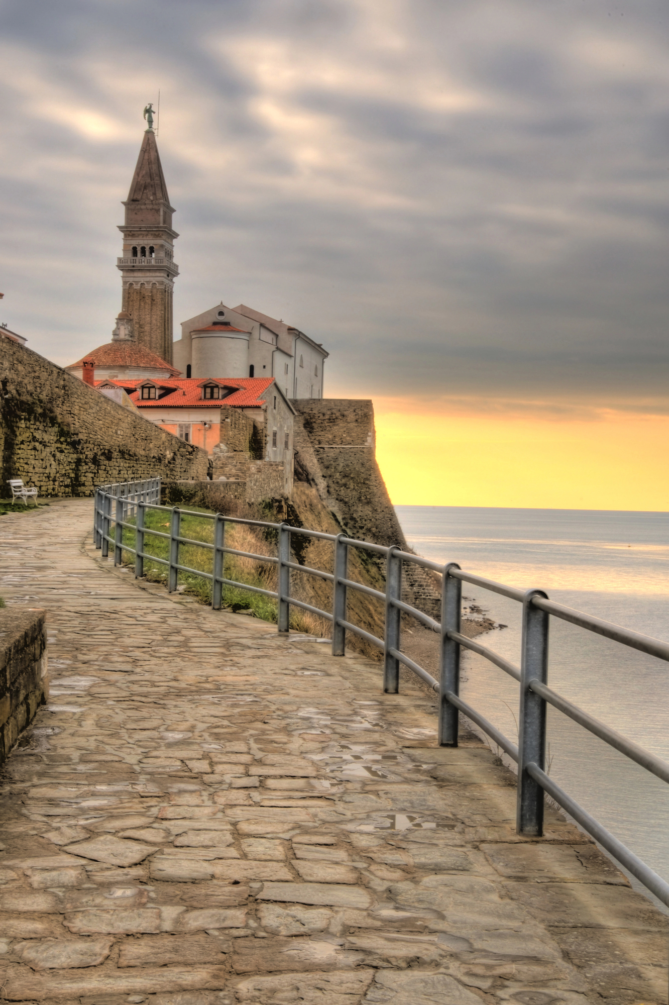 Stone pathway along a seaside cliff with a tower, old buildings, and a cloudy sunset sky&mdash;this charming scene offers the atmosphere of popular European destination dupes without the crowds.