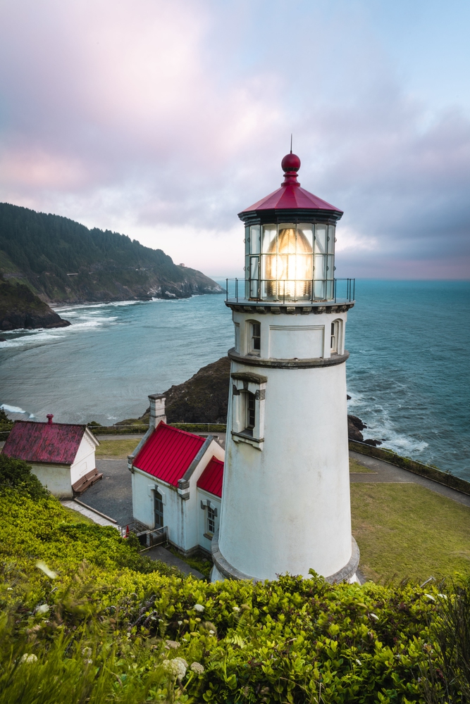 Lighthouse with a lit beacon on a cliff overlooking the ocean at sunset, red-roof buildings nearby&mdash;an idyllic scene reminiscent of hidden European destination dupes.