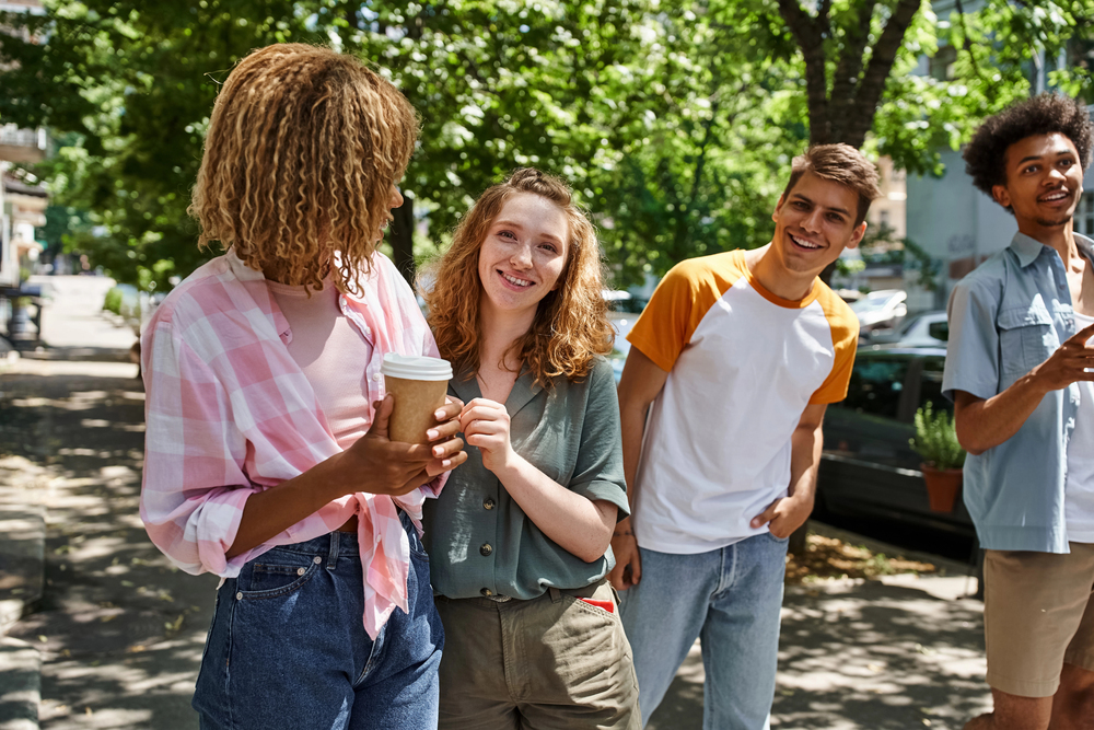 Four friends smile and chat outdoors on a sunny day, one holding a coffee cup, as they enjoy a break during their food tour. Trees and cars in the background complete the lively scene.