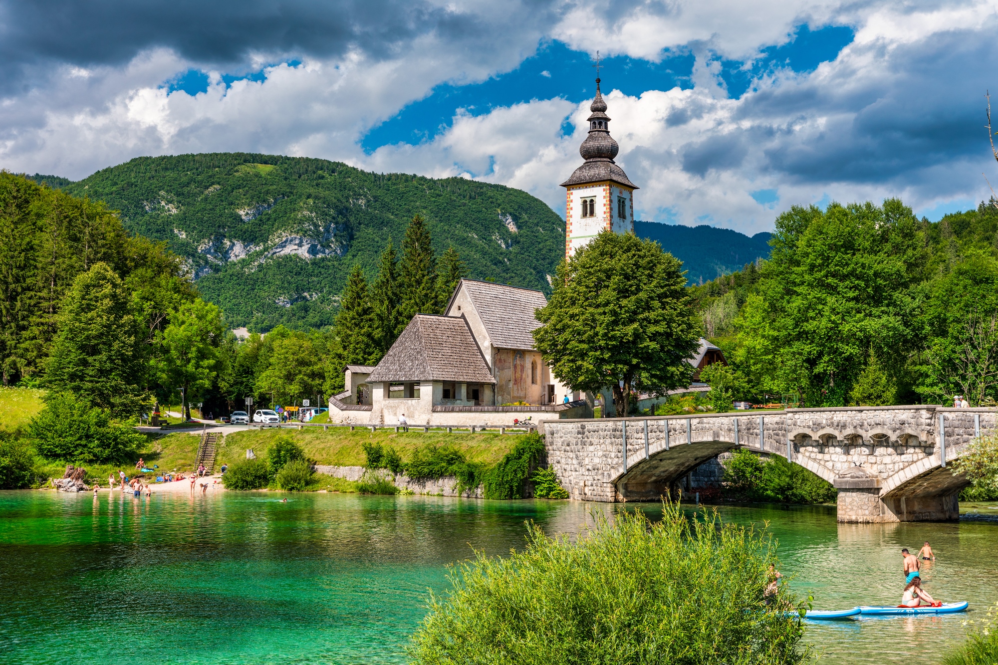 A stone bridge, church, and swimmers by a clear river with green hills and a cloudy sky in the background&mdash;this picturesque scene could be one of Europe&rsquo;s hidden gems, perfect for travelers seeking European destination dupes without the crowds.