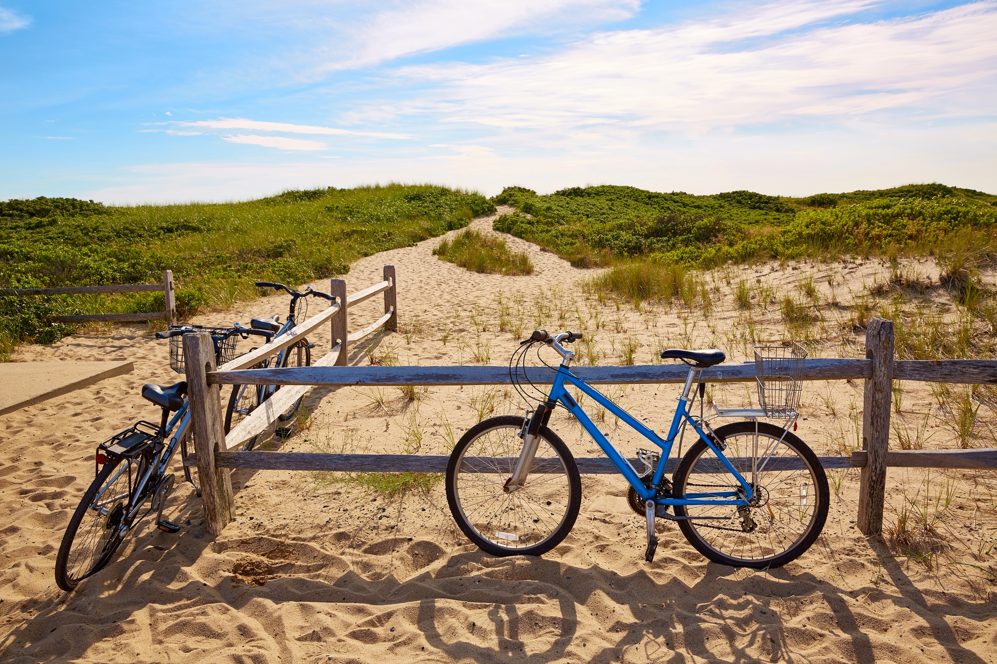 Two bicycles rest by a wooden fence near a sandy beach path through grassy dunes under a blue sky, capturing the serene charm often found in European destination dupes.
