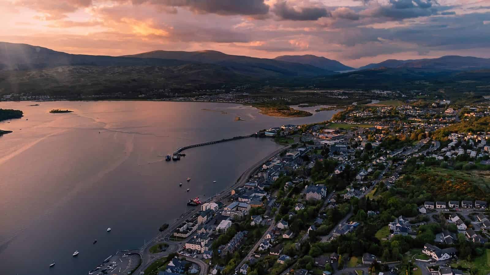 Aerial view of a coastal town at sunset with mountains, water, and scattered clouds in the sky.