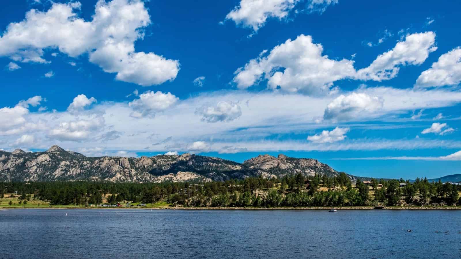Blue lake with pine-covered hills and rocky mountains under a bright blue sky with scattered white clouds.
