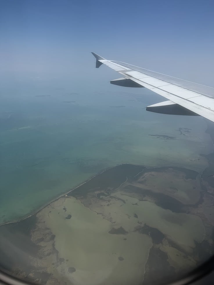 Aerial view of a coastline and ocean through an airplane window with a wing visible against a clear sky, reminiscent of the breathtaking landscapes described in The Rainforest Lodge at Sleeping Giant Review.