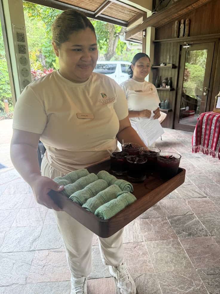 A woman serves a tray with rolled towels and drinks at The Rainforest Lodge at Sleeping Giant; another woman stands in the background holding papers.