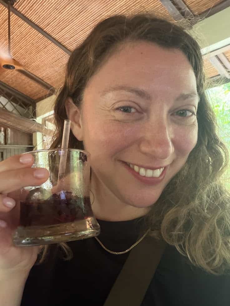 Smiling woman with wavy hair holds a drink with a straw, indoors near a window with natural light, reminiscent of the relaxed atmosphere detailed in The Rainforest Lodge at Sleeping Giant Review.