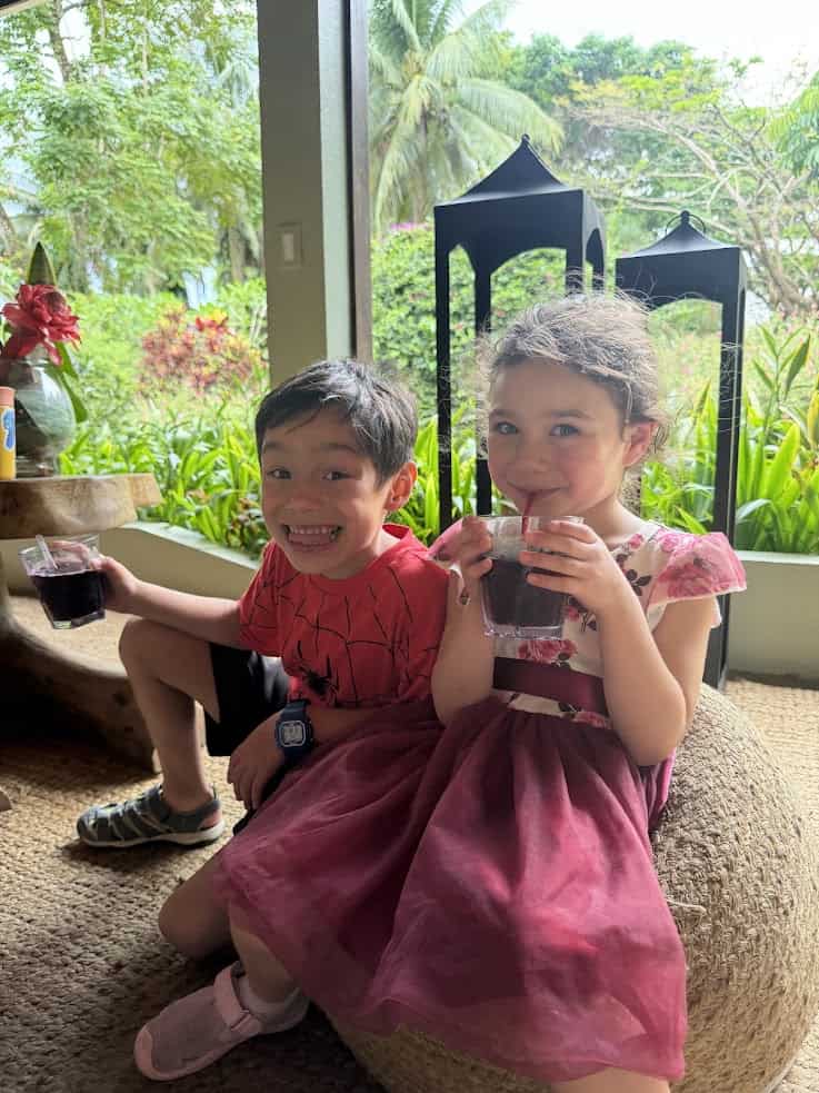Smiling boy and girl sitting together, drinking juice with straws, surrounded by lush greenery outdoors, reminiscent of a serene moment at The Rainforest Lodge at Sleeping Giant Review.