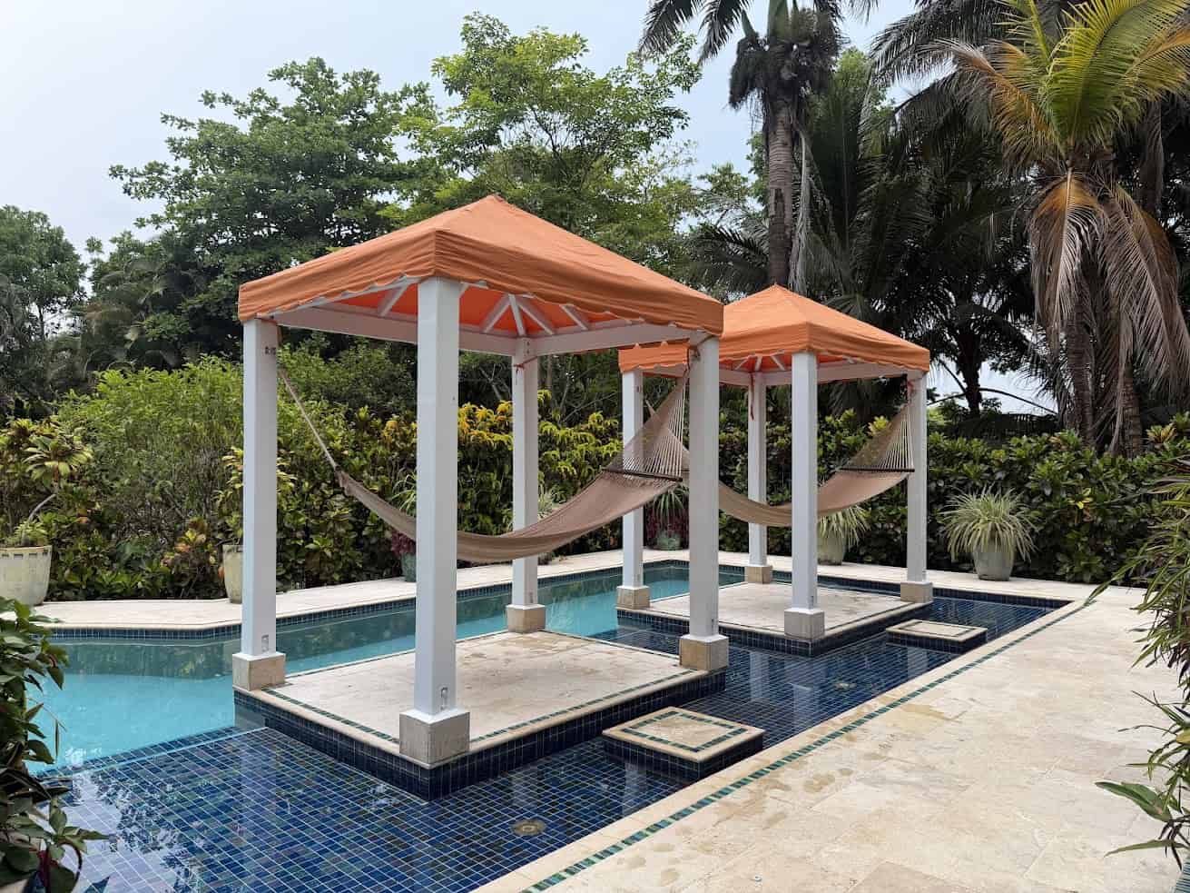 Two hammocks hang under orange canopies beside a tiled pool, surrounded by tropical plants and trees—an inviting scene reminiscent of the tranquil ambiance described in The Rainforest Lodge at Sleeping Giant Review.
