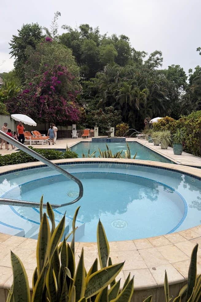 A round hot tub in front of a large pool, surrounded by plants, trees, and people relaxing nearby—just like the tranquil setting described in The Rainforest Lodge at Sleeping Giant Review.