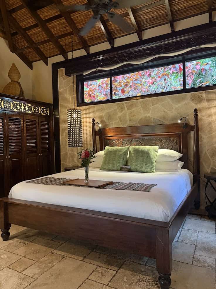 Wooden bed with green pillows, flowers on a tray, stone wall, and plants visible through a window evoke the serene ambiance of The Rainforest Lodge at Sleeping Giant Review.