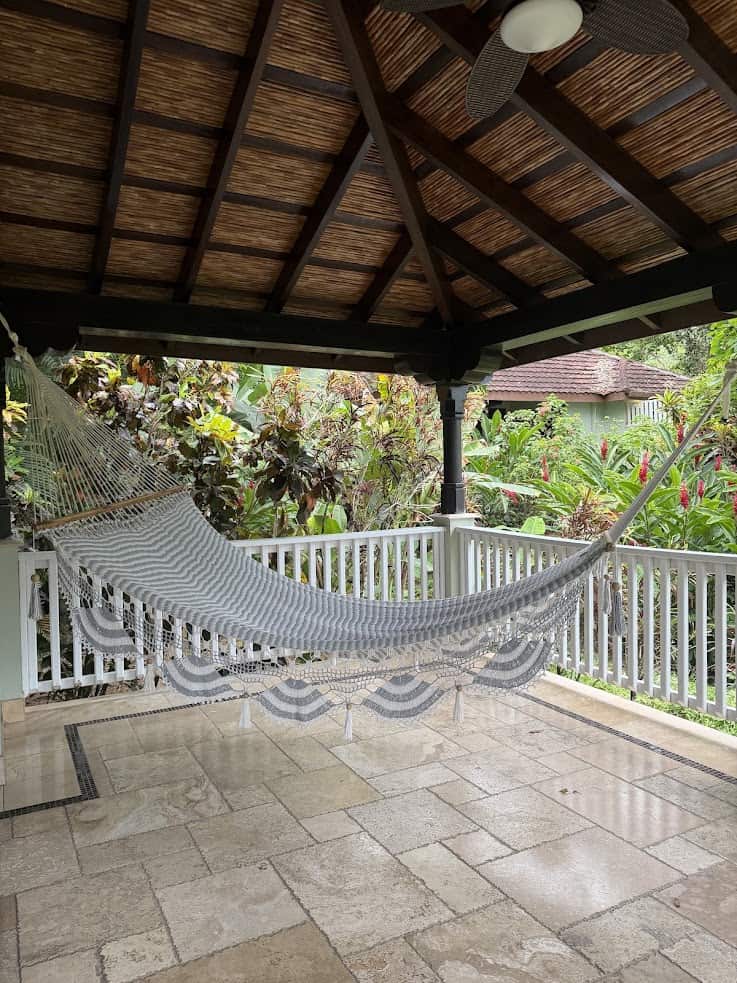 A hammock hangs in a covered patio with tiled flooring, white railing, and lush plants in the background, evoking the tranquil charm praised in The Rainforest Lodge at Sleeping Giant Review.