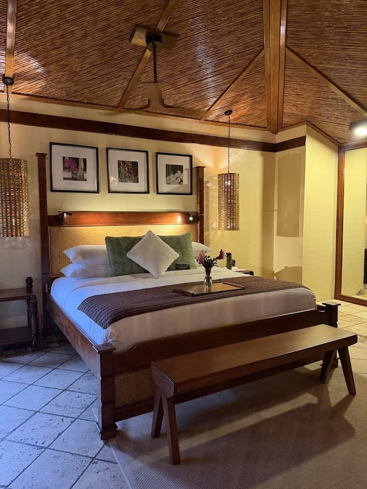 A cozy hotel room at The Rainforest Lodge at Sleeping Giant features a wooden bed, green and white pillows, artwork, and soft lighting.