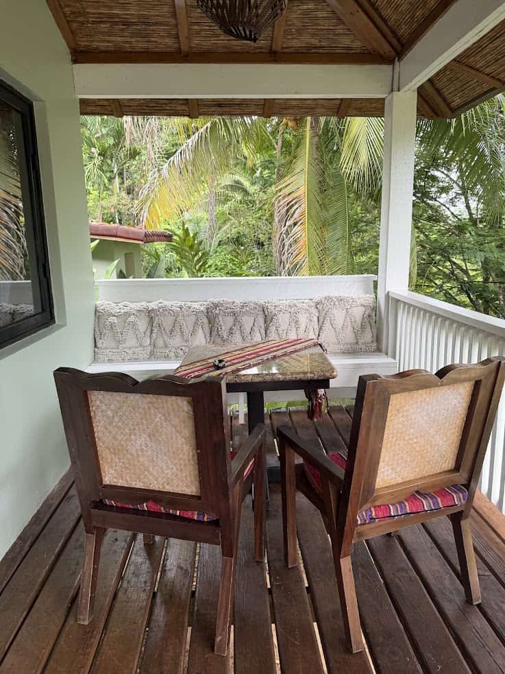 A cozy porch with two wooden chairs, a table, and a cushioned bench, surrounded by greenery, evokes the tranquil atmosphere described in The Rainforest Lodge at Sleeping Giant Review.