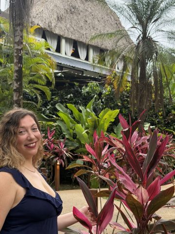 Woman smiling near red leafy plants, with lush greenery and a thatched-roof building in the background—an inviting scene reminiscent of The Rainforest Lodge at Sleeping Giant Review.