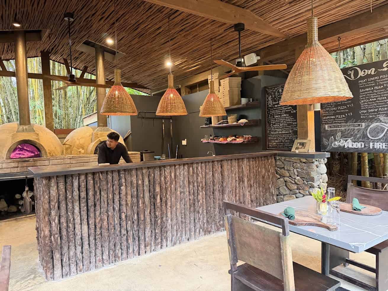 Outdoor wood-fired pizza kitchen with a chef behind the counter, stone oven, and menu boards on the wall—reminiscent of the rustic charm you’d find in The Rainforest Lodge at Sleeping Giant Review.