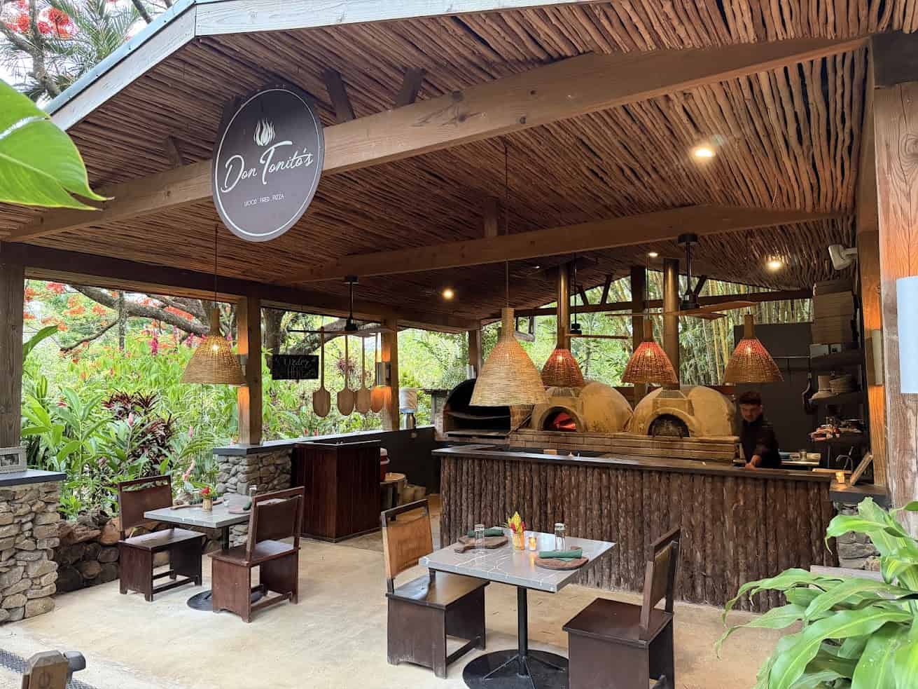Outdoor restaurant with wood-fired ovens, wooden tables, hanging lights, and lush green surroundings—an experience reminiscent of The Rainforest Lodge at Sleeping Giant Review.