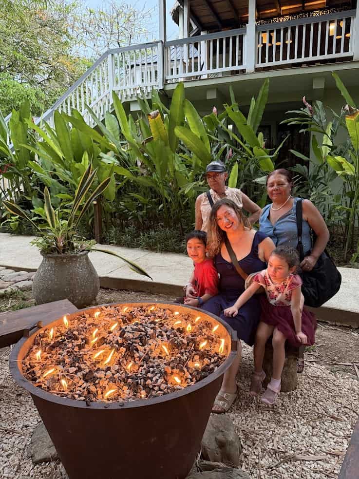 Five people sit and stand near a large outdoor fire pit, surrounded by lush plants and a white house, evoking the tranquil ambiance often described in The Rainforest Lodge at Sleeping Giant Review.