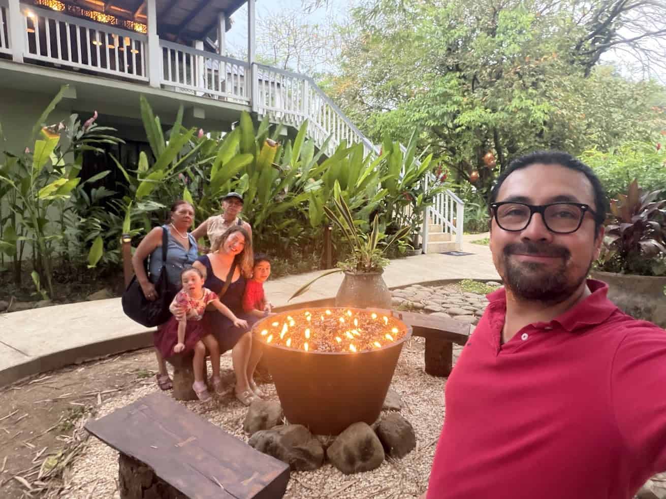 A group of people smiles by a fire pit in a lush garden, with a house in the background, reminiscent of the warm and welcoming atmosphere highlighted in The Rainforest Lodge at Sleeping Giant Review.