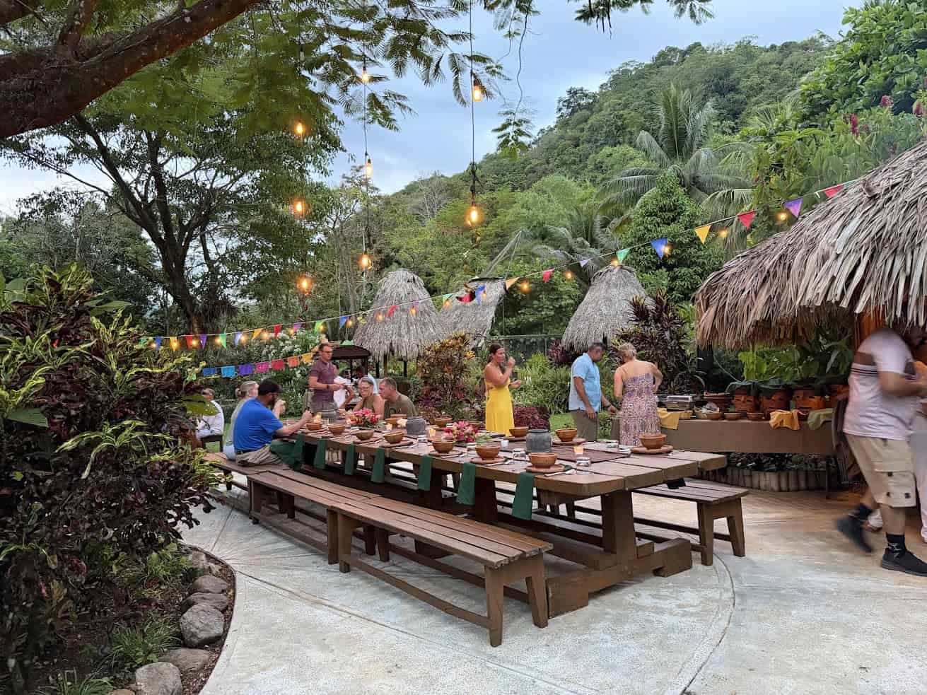 Outdoor tropical party with people eating at long tables, festive lights, huts, and colorful pennant banners—an atmosphere reminiscent of The Rainforest Lodge at Sleeping Giant Review.
