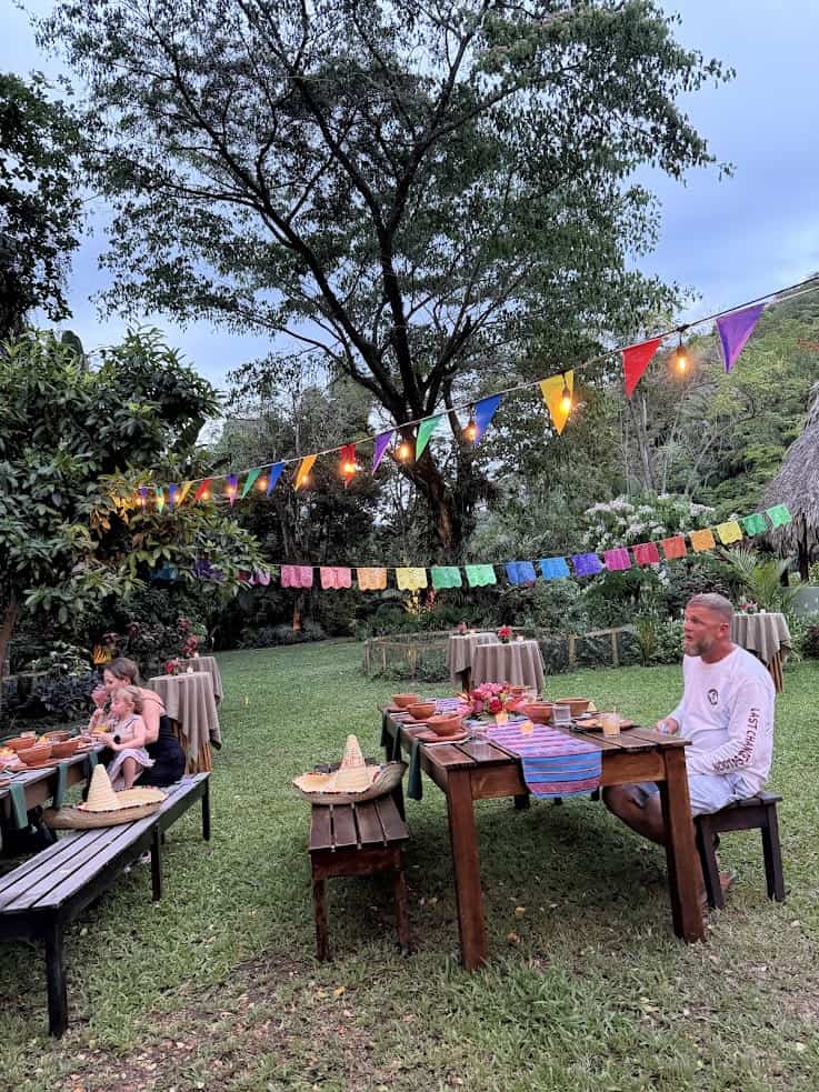 Outdoor picnic with colorful flags, people sitting at tables, trees, and string lights in the background—an inviting setting reminiscent of a scene you might find in a The Rainforest Lodge at Sleeping Giant Review.