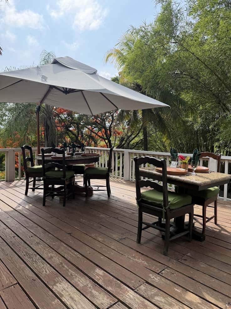 Wooden patio with tables, chairs, and a large umbrella, surrounded by lush green trees and flowers—an inviting outdoor space reminiscent of tranquil settings found in many The Rainforest Lodge at Sleeping Giant reviews.