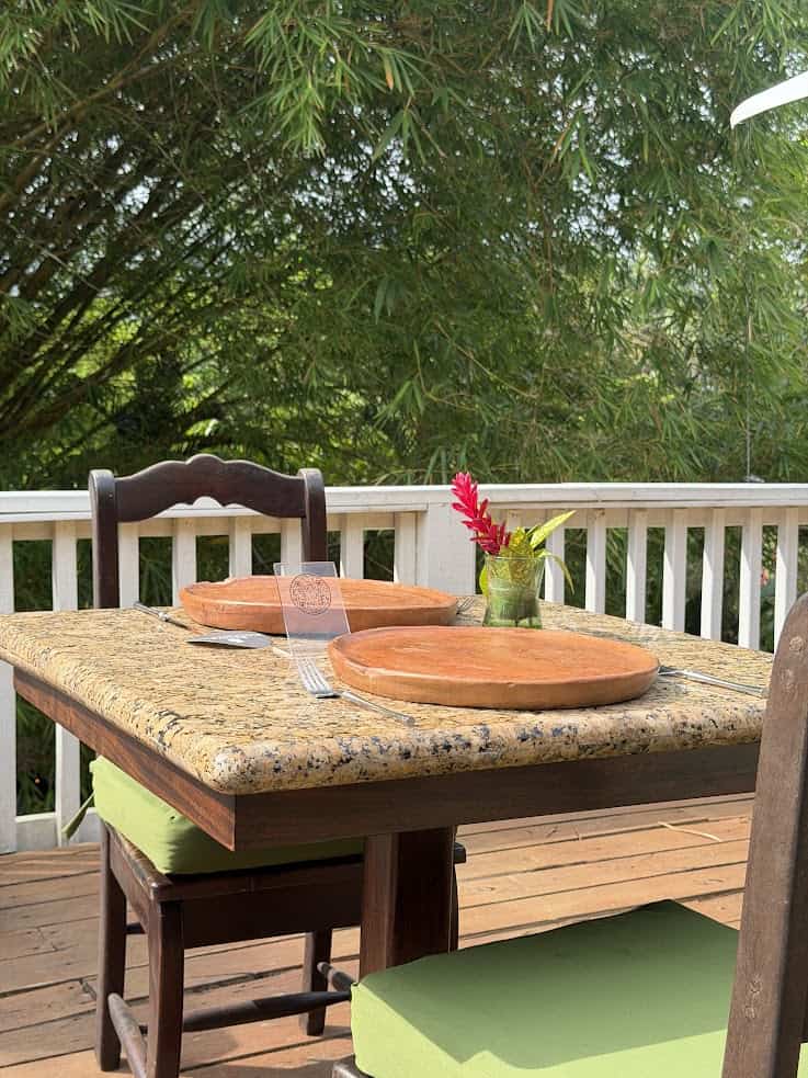 Granite table with two wooden chairs, plates, and a vase of flowers on a deck with greenery behind—a serene setting reminiscent of the tranquil ambiance described in The Rainforest Lodge at Sleeping Giant Review.