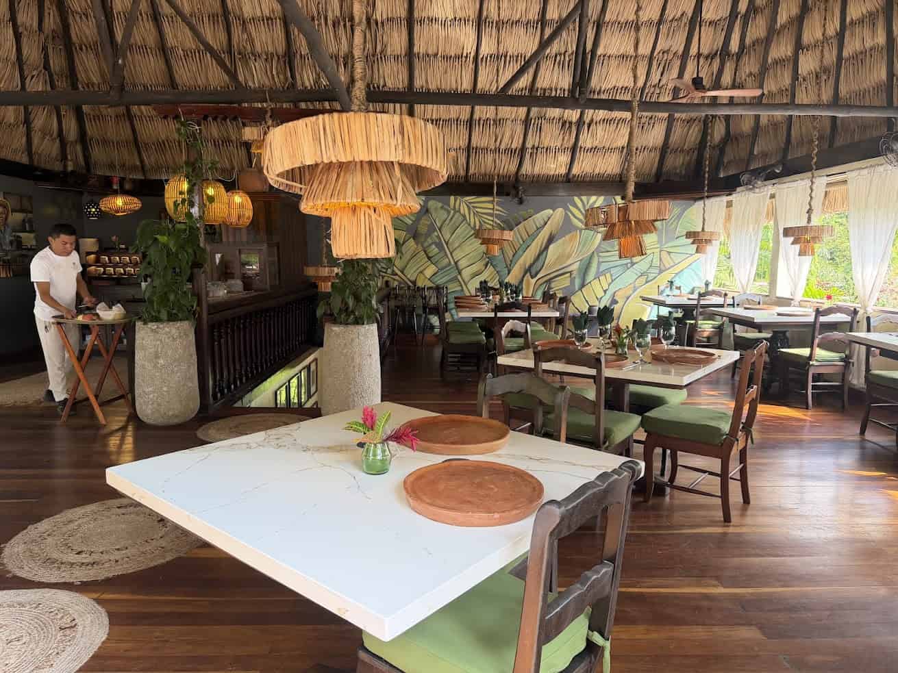 Chic restaurant with thatched roof, leafy mural, wooden tables, green chairs, and natural light—evoking vibes reminiscent of The Rainforest Lodge at Sleeping Giant Review.