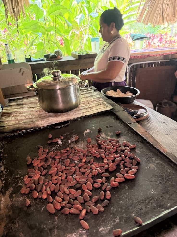 Cocoa beans roasting on a griddle with a woman cooking in a rustic kitchen in the background, reminiscent of the authentic culinary experiences highlighted in The Rainforest Lodge at Sleeping Giant Review.