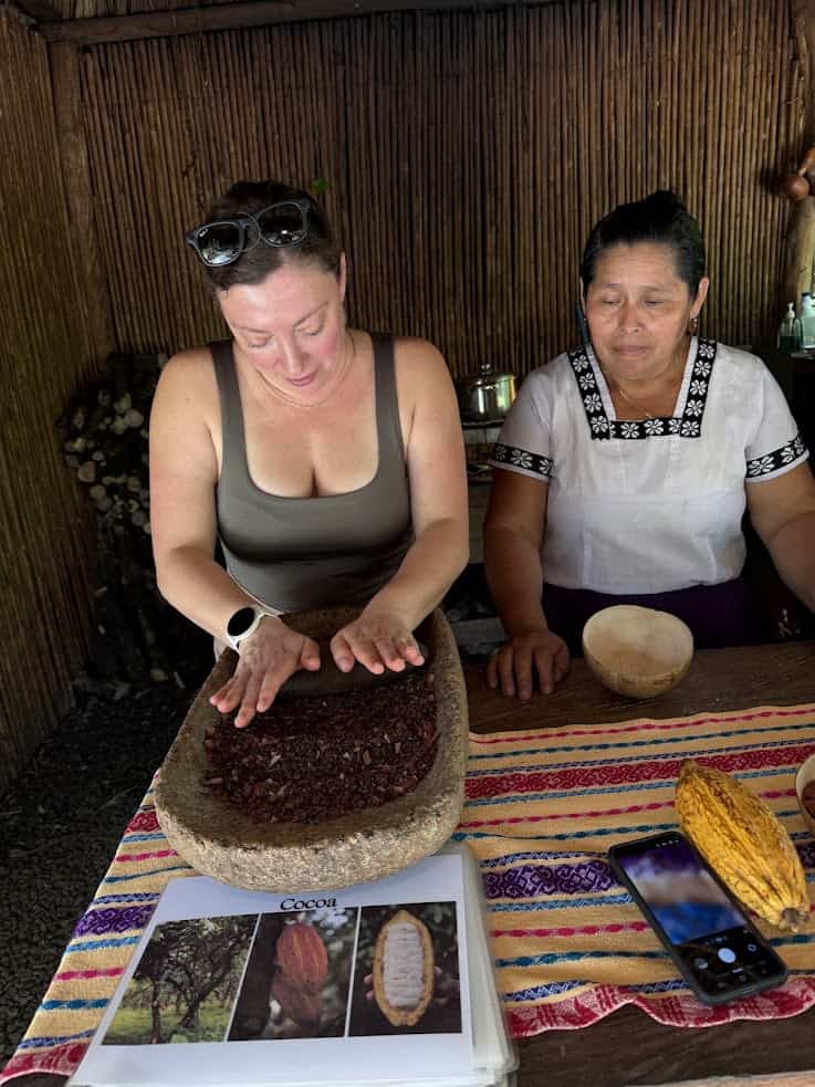 Two women prepare cacao on a stone table at The Rainforest Lodge at Sleeping Giant; cacao pods and a booklet are also on the striped cloth.