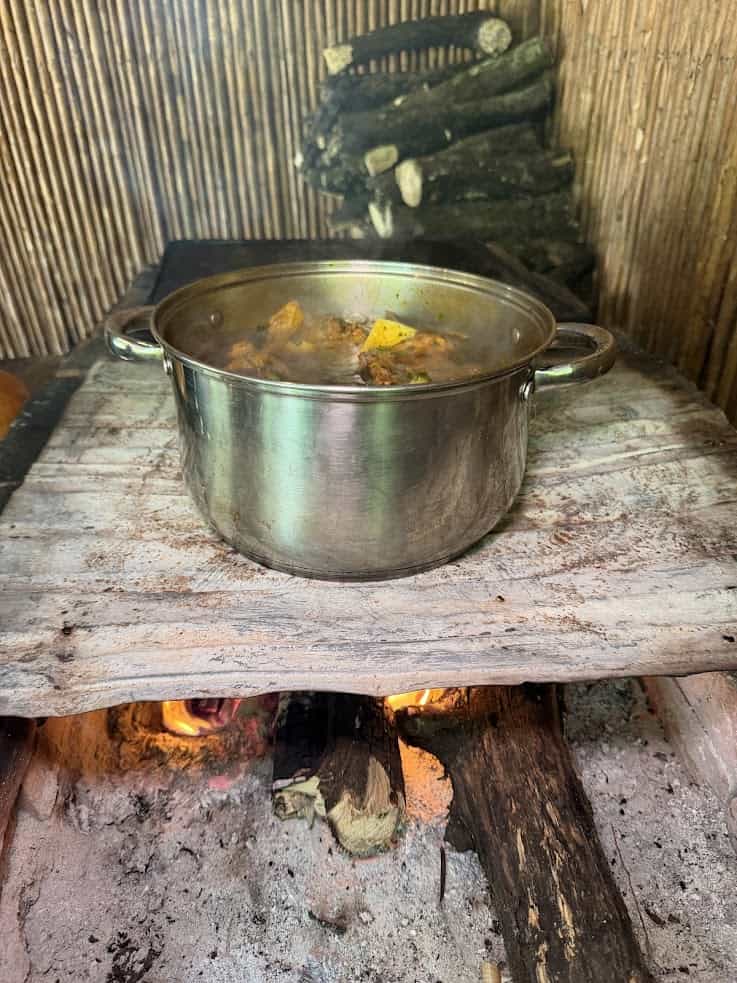 A metal pot with food cooking over an open wood fire on a rustic stove with logs and wooden walls, reminiscent of the authentic culinary experiences described in The Rainforest Lodge at Sleeping Giant Review.