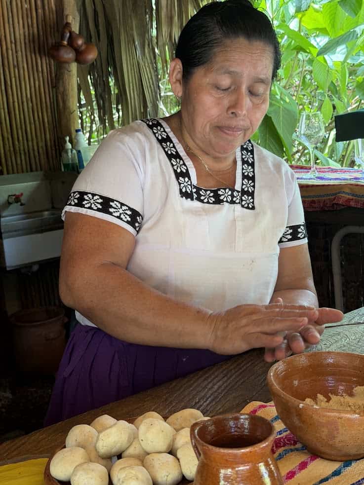 Woman in traditional dress shapes dough balls by hand at a rustic kitchen counter with pottery and plants, evoking the homely atmosphere described in The Rainforest Lodge at Sleeping Giant Review.