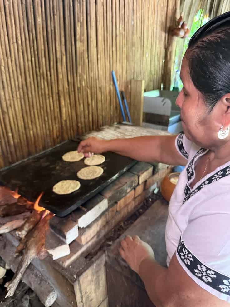 A woman cooks tortillas on a wood-fired stove in a rustic kitchen with bamboo walls, capturing the authentic atmosphere you might find in The Rainforest Lodge at Sleeping Giant—reviewing traditional culinary experiences in an enchanting jungle setting.