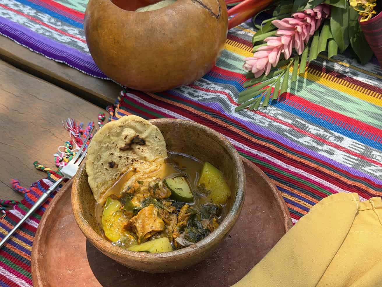 A bowl of stew with vegetables and a tortilla on top, set on a colorful woven cloth with flowers nearby, evokes the vibrant dining experiences described in The Rainforest Lodge at Sleeping Giant Review.