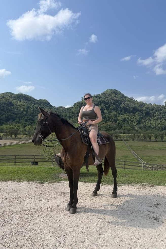 Woman in sunglasses riding a dark horse on a ranch with green hills and blue sky in the background, reminiscent of the scenic vistas featured in The Rainforest Lodge at Sleeping Giant Review.