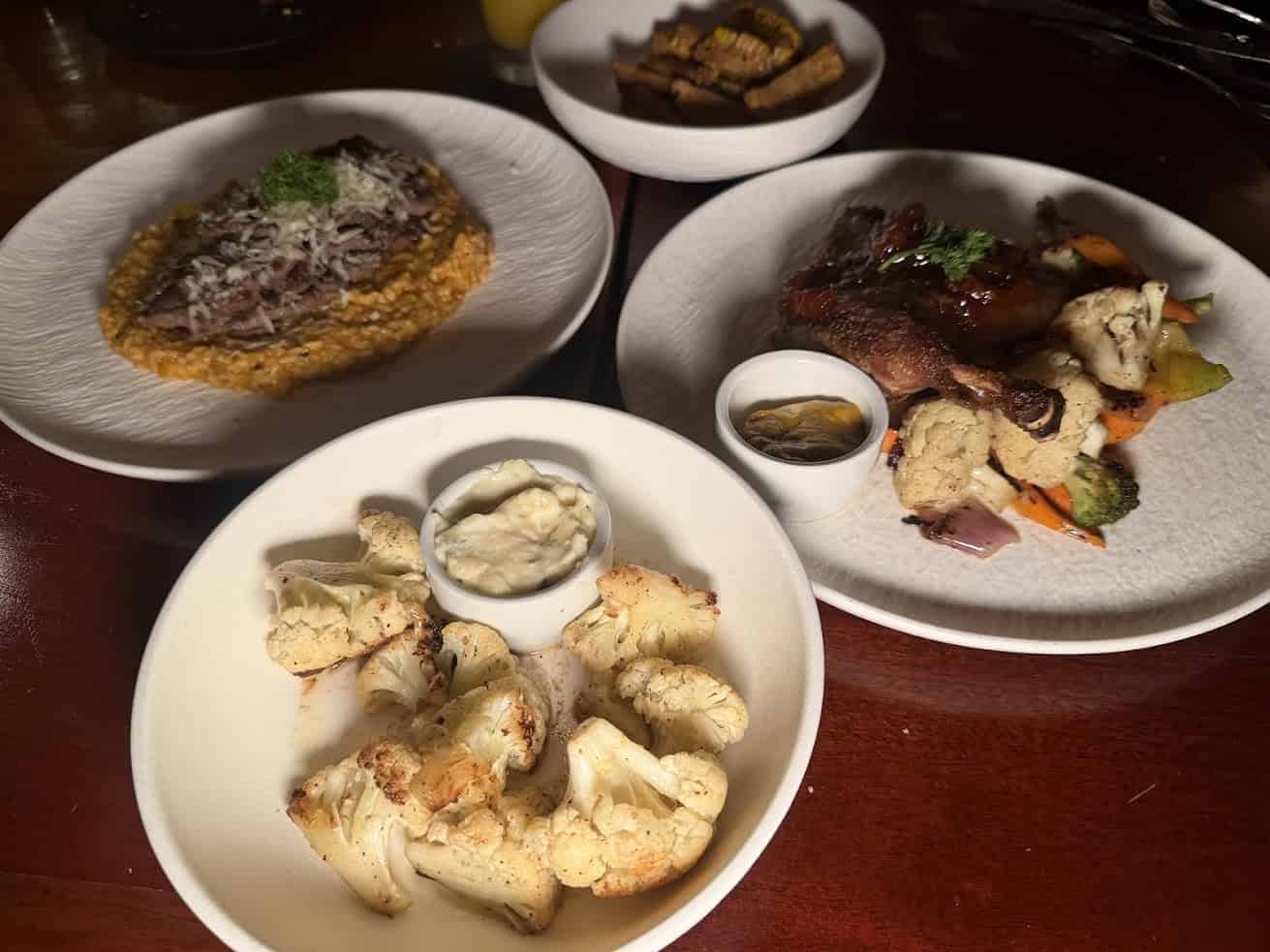 Four plates with roasted cauliflower, grilled chicken and vegetables, risotto, and a small dish of dip are beautifully arranged on a table—an inviting spread reminiscent of the culinary delights highlighted in The Rainforest Lodge at Sleeping Giant Review.