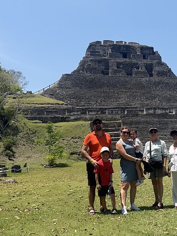 Six people stand in front of a large ancient stone pyramid surrounded by grass and trees on a sunny day, enjoying a memorable stop on their Belize reef and ruins trip.