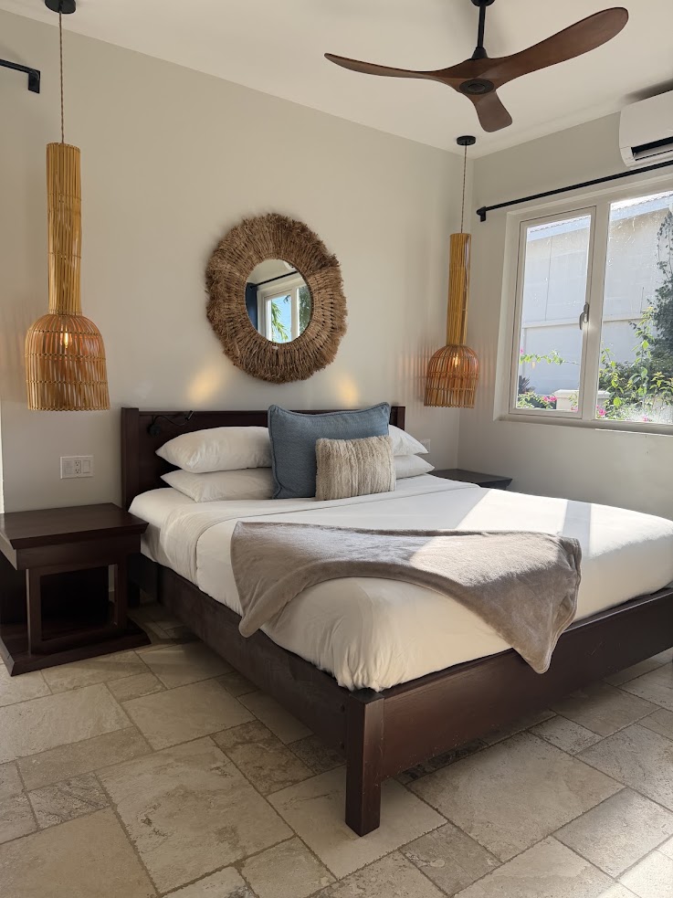 Sunlit bedroom with a neatly made bed, wicker lamps, a round mirror, and a window with garden view&mdash;this inviting setting evokes the serene comfort praised in many a The Lodge at Jaguar Reef review.