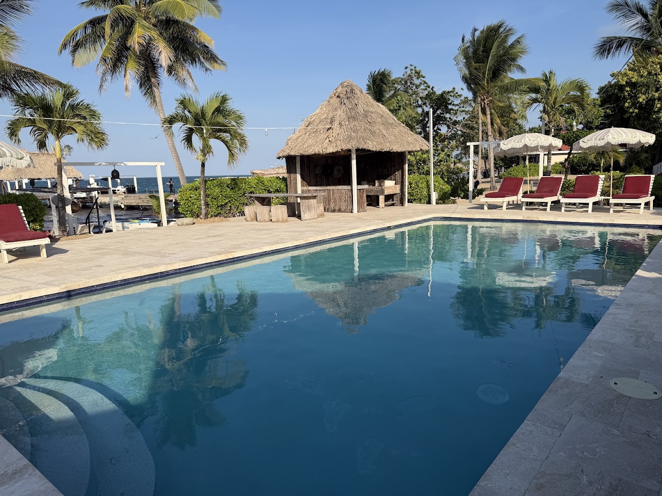 A clear swimming pool with red lounge chairs, palm trees, and a thatched hut under a blue sky offers the ultimate tropical retreat&mdash;just like those described in every glowing The Lodge at Jaguar Reef review.
