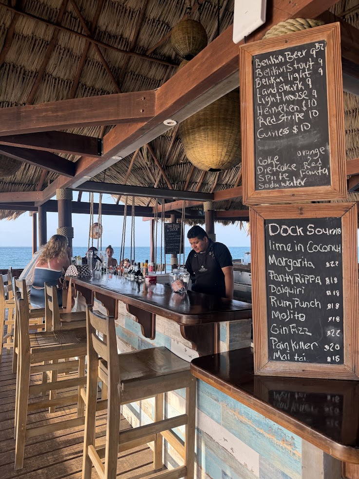 Open-air beach bar with a thatched roof, wooden stools, and handwritten drink menus by the counter&mdash;a setting that echoes the laid-back ambiance praised in every The Lodge at Jaguar Reef review.