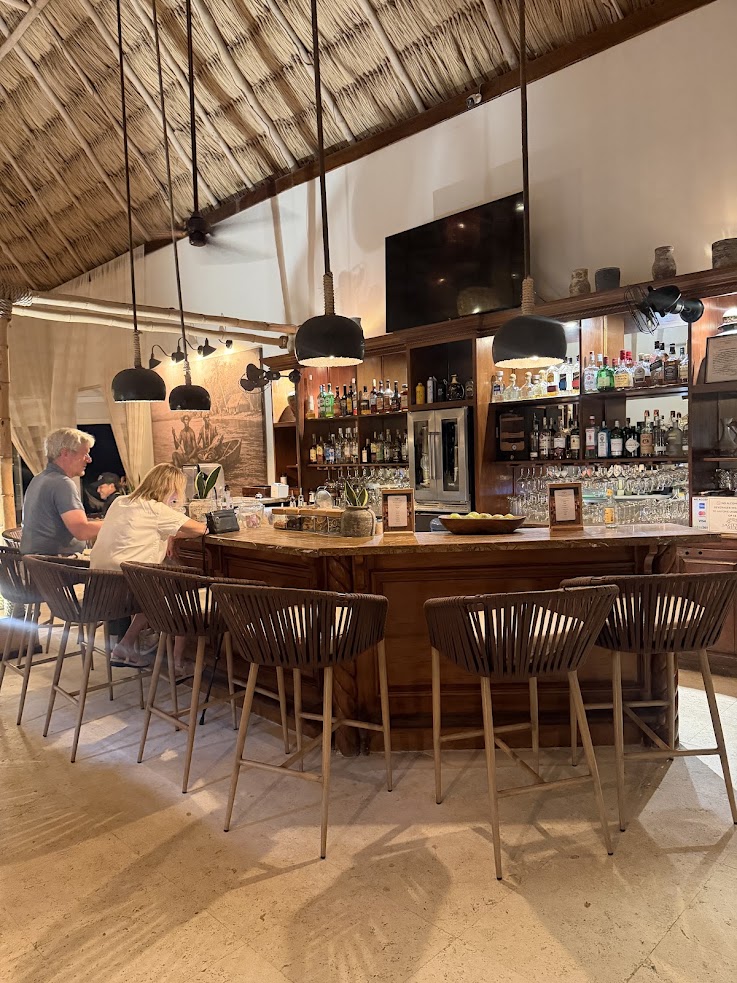 A cozy bar with wooden stools, people sitting, and bottles lined up behind the counter under a thatched roof, just like the inviting atmosphere mentioned in many a The Lodge at Jaguar Reef review.