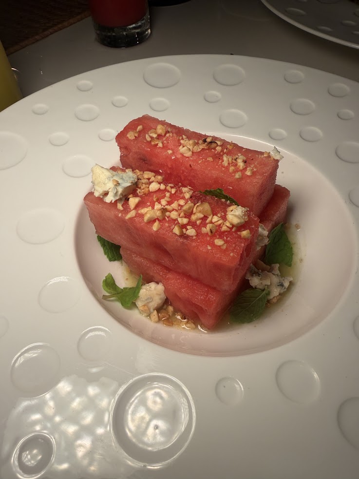 Stacked watermelon slices topped with crumbled cheese, chopped nuts, and mint on a white plate&mdash;an appetizer reminiscent of the fresh, vibrant flavors often highlighted in The Lodge at Jaguar Reef review.