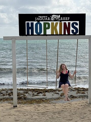 A person sits on a swing by the beach under a sign that reads “JAGUAR REEF HOPKINS,” capturing a tranquil moment that perfectly reflects the serene beauty often praised in the lodge at jaguar reef review.