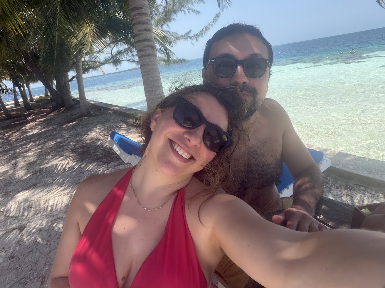 Smiling couple in sunglasses take a selfie on a sunny beach with clear water and palm trees, capturing the perfect moment for their The Lodge at Jaguar Reef review.