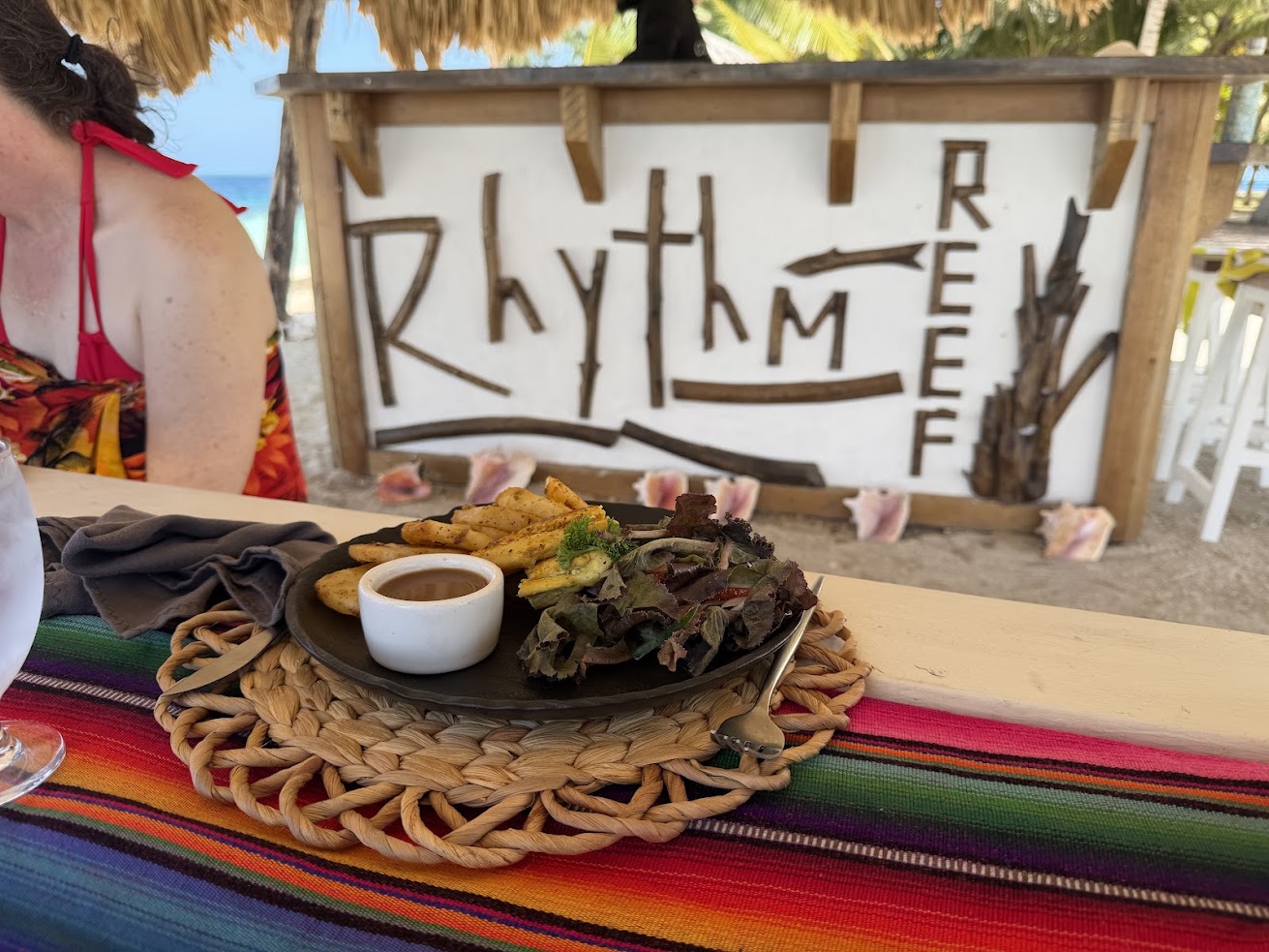 Plate of food on a colorful tablecloth with a &ldquo;Rhythm Reef&rdquo; sign and beach d&eacute;cor in the background, capturing the vibrant ambiance reminiscent of experiences highlighted in many The Lodge at Jaguar Reef reviews.