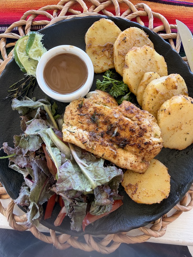 Grilled chicken breast with sliced potatoes, salad greens, lime wedge, and dipping sauce on a black plate&mdash;an inviting dish reminiscent of those featured in a glowing The Lodge at Jaguar Reef review.