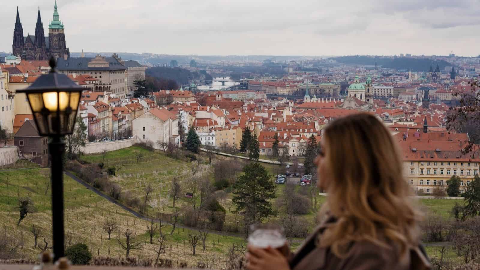 A woman with a drink overlooks Prague’s red rooftops and Prague Castle on a cloudy day.
