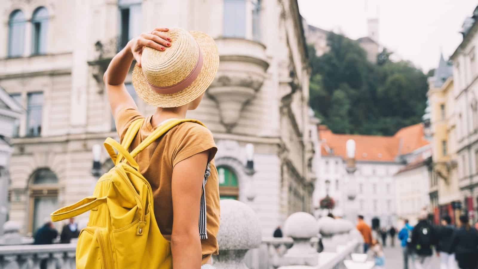 Person with a yellow backpack and hat looks at historic city buildings, standing on a stone bridge.