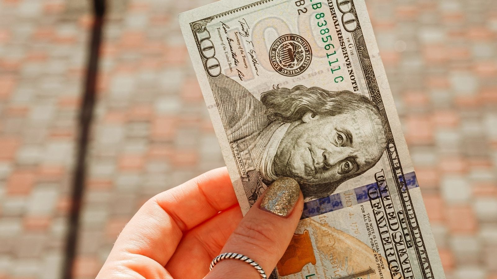 Hand with painted nails holding a $100 bill against a blurred, checkered ground background.
