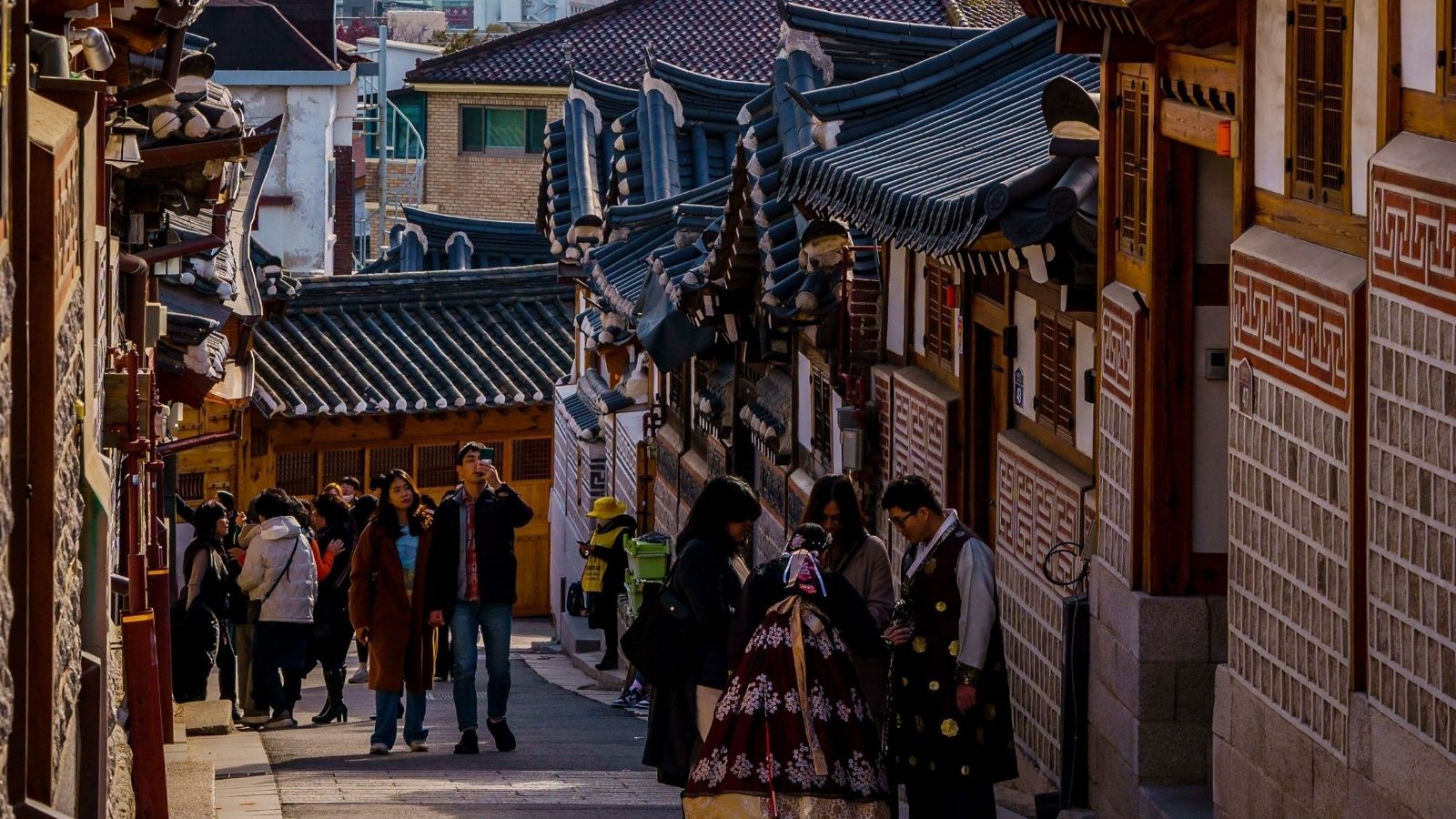 People walk along a street lined with traditional Korean houses on a sunny day.