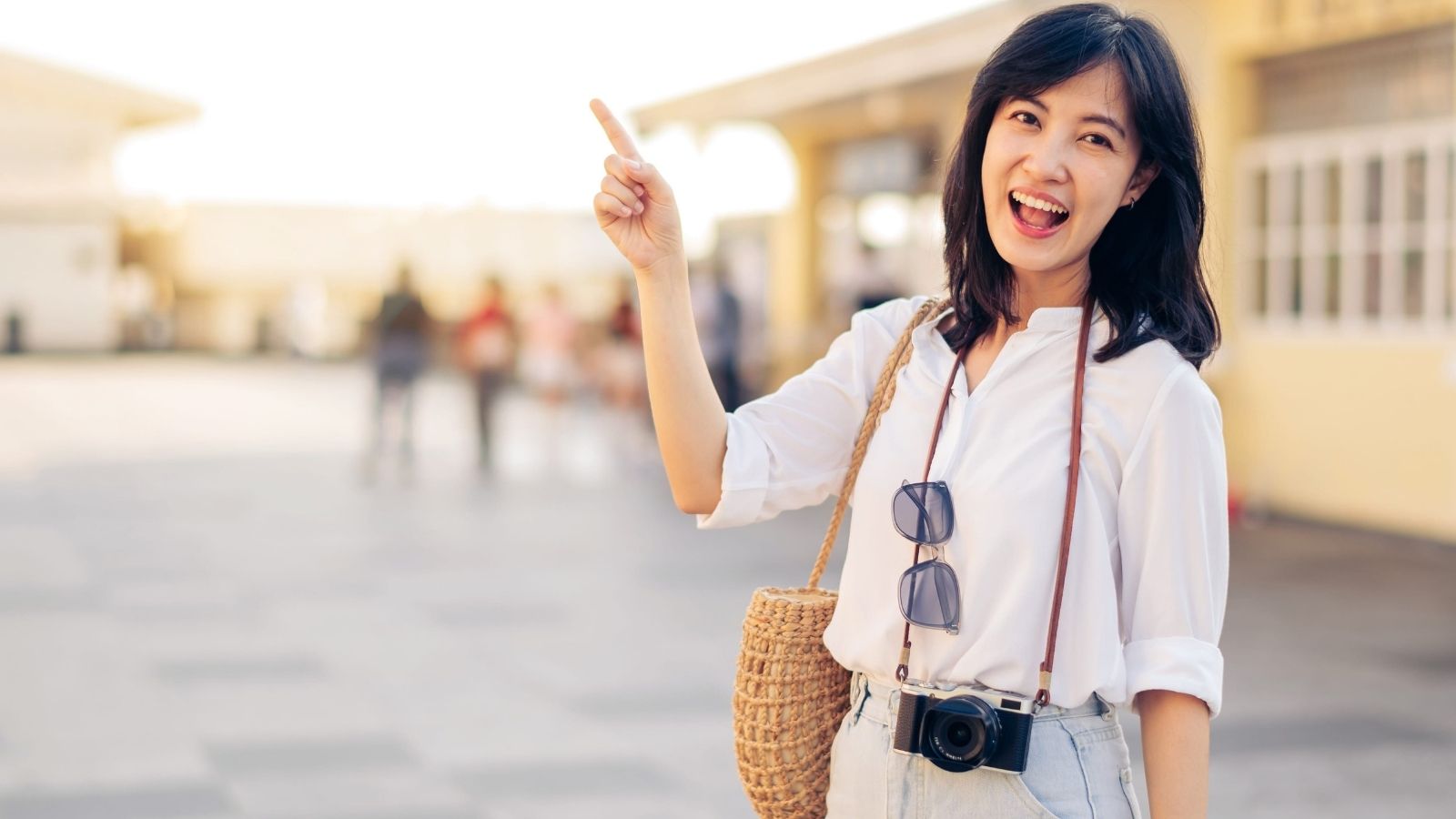Woman in white shirt smiling, pointing up, with camera, sunglasses, and woven bag outdoors.